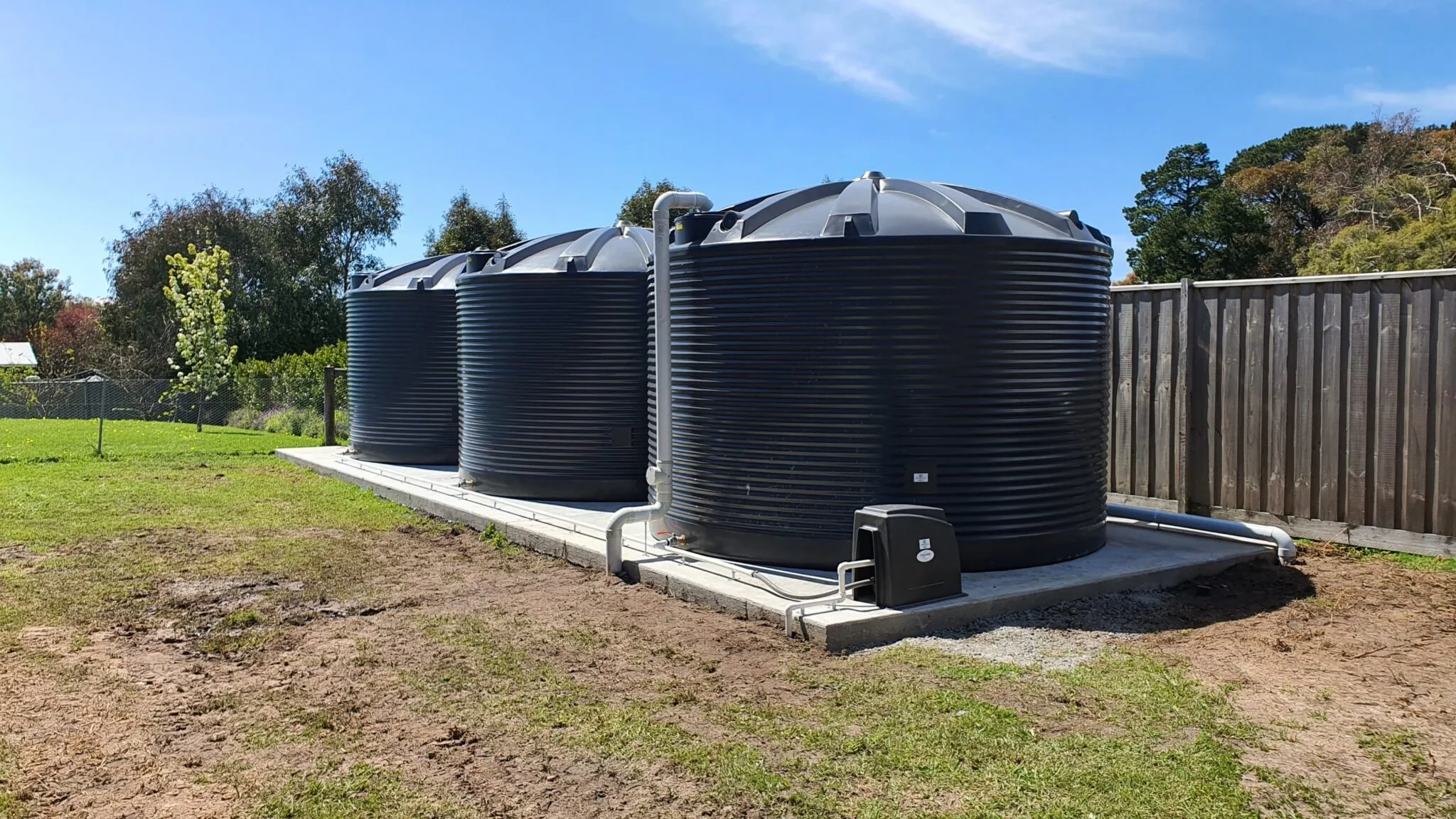 Three Polymaster round rainwater tanks on a concrete base with a water pump system installed, set in a residential backyard